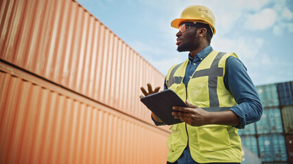 Smiling Portrait of a Handsome African American Black Industrial Engineer in Yellow Hard Hat and Safety Vest Working on Tablet Computer. Inspector or Safety Supervisor in Container Terminal.