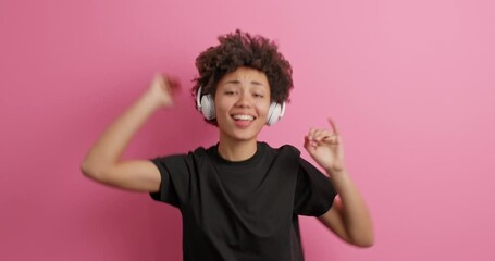 Wall Mural - Joyful curly haired woman jumps and dances catches every bit of music wears casual black t shirt enjoys song in wireless headphones isolated over pink background. Amused entertained teenage girl