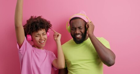 Poster - Positive carefree dark skinned ethnic couple dance together while listen favorite music via wireless headphones move with rhytm of melody isolated over pink background. People and lifestyle concept