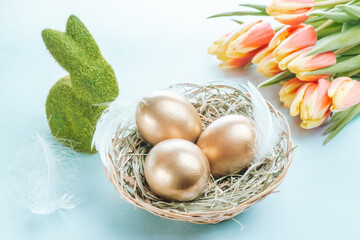 Wall Mural - Golden easter colour eggs in basket with spring tulips, white feathers on pastel blue background in Happy Easter decoration. Spring holiday concept.