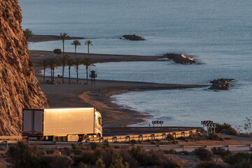 Canvas Print - Truck with refrigerated semi-trailer on a mountain road that goes down to a beach with palm trees.