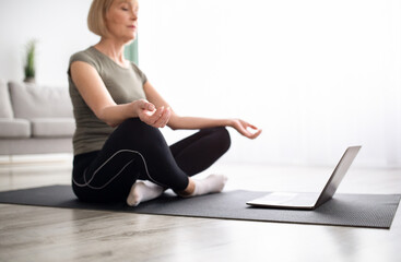 Wall Mural - Balanced mature woman meditating with closed eyes in front of laptop at home, selective focus