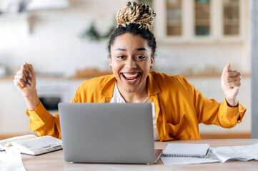 Wall Mural - Happy beautiful successful young African American woman with dreadlocks, manager, freelance, agent, works at home using laptop, rejoice at victory or successful deal, gesturing with hands, smiling