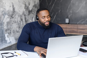 Unshaved black businessman making video conference on laptop sitting at home, wearing black headset, professional call center agent consulting, man working at office and talking, quarantine concept.