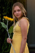 Vertical outdoor portrait of a beautiful tween girl with long blonde hair wearing a yellow dress, holding a sunflower and smiling at the camera