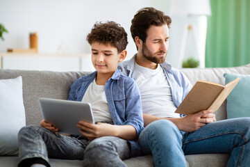 Wall Mural - No communication in family. Father reading book while his son using digital tablet, sitting together on sofa