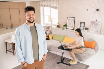 Poster - Young cheerful man in casualwear looking at you while standing against his wife sitting on couch and using laptop while networking