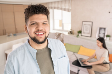 Poster - Happy young bearded man in casualwear looking at you with toothy smile against his wife sitting on couch and using laptop in living-room
