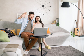 Poster - Young cheerful couple in casualwear looking at their friends on laptop display with toothy smiles and waving hands in home environment