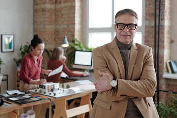 Sticker - Smiling mature male architect in smart casualwear and eyeglasses standing against his two young colleagues working over model of new house