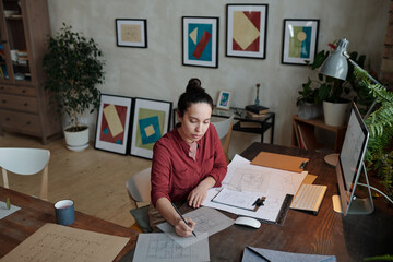 Poster - Young serious female architect with pencil drawing sketch of new construction while sitting by table in front of computer monitor