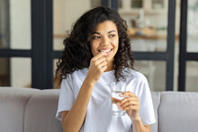 Young Beautiful Smiling African American Woman Holding  Holding Vitamin Pill And Glass Of Water Sitting  On The Couch At Home. Healthy Lifestyle, Healthy Diet Nutrition Concept