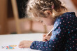 © Aleksandr - Close up of little girl draw in the album using color pensins. Cute girl paints coloring page at home during quararntine. Happy kid sits at the table by the window