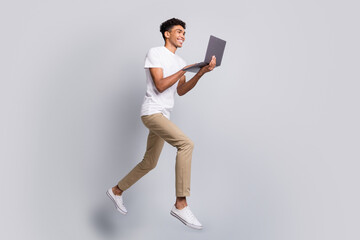 Poster - Full size profile photo of brunet optimistic curly guy jump hold laptop wear white t-shirt pants isolated on grey background