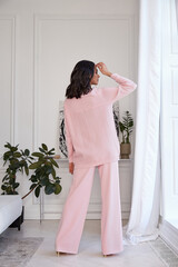 Wall Mural - Pretty fashion beautiful woman sexy lady brunette curly hair dark tanned skin wear trend clothes knitted pink pink suit jacket top pants shoes interior room sofa plants spring collection luxury life.