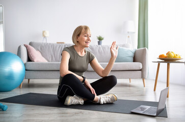 Wall Mural - Mature woman in sports clothes sitting on yoga mat in front of laptop, ready for domestic workout, greeting her trainer
