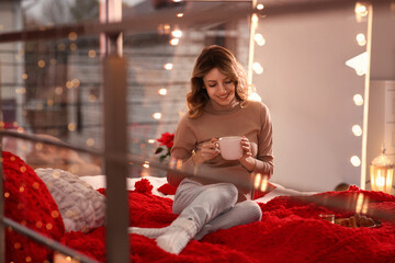 Sticker - Beautiful young woman with cup of drink relaxing on bed at home. Cozy atmosphere