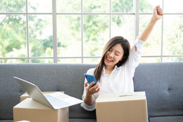 Happy young Asian woman entrepreneur, Smile for sales success after checking order from online shopping store in a smartphone at home office, Concept of merchant business online and eCommerce