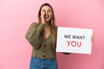Wall Mural - Young caucasian woman isolated on pink background holding We Want You board and shouting