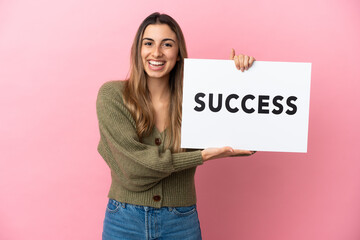 Wall Mural - Young caucasian woman isolated on pink background holding a placard with text SUCCESS with happy expression