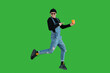 Cheerful guy in stylish casual clothes jumps with a eco cup in his hand and points a finger at it on a green background and looks at the camera with a smile on his face.