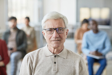 Portrait of white haired senior man smiling at camera while posing in office setting with people in background, copy space