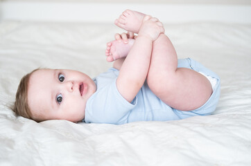 funny 6 months baby boy playing with legs, lying on bed at home on white bedding.