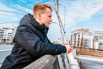 A young blond man in a jacket stands on the pedestrian part of a futuristic modern bridge with beams in St. Petersburg