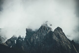 Fototapeta Góry - Dark atmospheric surreal landscape with dark rocky mountain top in low clouds in gray cloudy sky. Gray low cloud on high pinnacle. High black rock with snow in low clouds. Surrealist gloomy mountains.