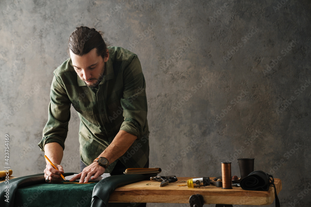 Bearded focused craftsman working with leather while standing at table - obrazy, fototapety, plakaty 