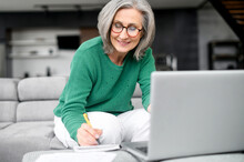 Confident Mature Woman Using Laptop Computer For Remote Work, Watching Webinar And Taking Notes Sitting At Home. Contemporary Senior Female Online Teacher Holding Video Conference And Making Marks