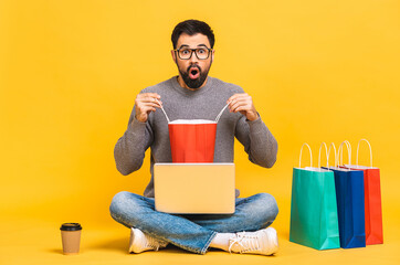 Wall Mural - Shopping time! Portrait of happy young bearded man with shopping packages holding laptop computer while sitting on floor isolated on yellow background.