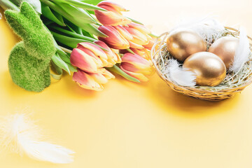 Wall Mural - Basket easter decoration: Golden eggs in basket with spring tulips, white feathers on pastel yellow background. Congratulatory easter design.