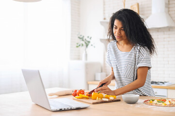 Focused mixed-race woman with afro hairstyle watching cooking classes, learning how to make yummy pizza dinner lunch watching video blog course from laptop in the modern kitchen, chopping veggies