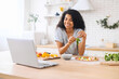 Mixed-race girl with afro hairstyle watching cooking classes, learning how to make yummy pizza dinner lunch watching video blog course from laptop in the modern kitchen, smiling, holding basil leaf