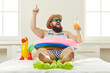Funny guy in sunglasses, sun hat and swim ring sitting on beach mattress and sipping cocktail points finger up struck by cool idea on how to turn quarantine holiday at home into fun summer vacation