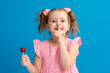 © skif - Close-up of a funny child showing a finger gesture to his lips. naughty little girl with a lollipop on a blue background in the studio posing with pleasure. Copy the space.