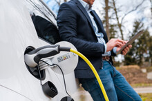 Business Man Standing Near Charging Electric Car Or EV Car And Using Tablet In The Street. 