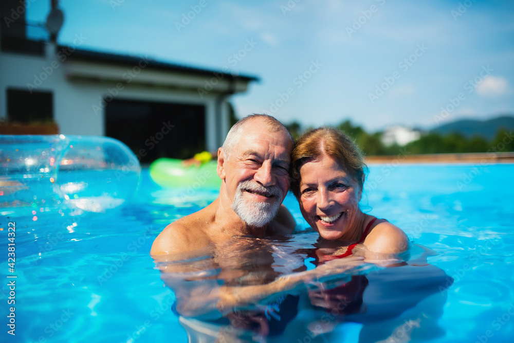 Cheerful senior couple in swimming pool outdoors in backyard, looking at camera. - obrazy, fototapety, plakaty 
