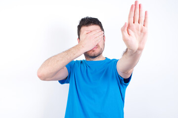 Wall Mural - young handsome caucasian man wearing blue t-shirt against white background covers eyes with palm and doing stop gesture, tries to hide from everybody.