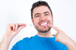 © Roquillo - young handsome caucasian man wearing blue t-shirt against white background holding an invisible aligner and pointing to her perfect straight teeth. Dental healthcare and confidence concept.