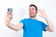 © Roquillo - young handsome caucasian man wearing white t-shirt against white background holds modern mobile phone and makes video call waves palm in hello gesture. People modern technology concept