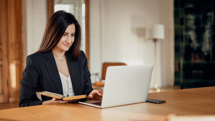 Wall Mural - The assistant works on a laptop computer in the office. Comfortable working space in a coworking space. Portrait of a beautiful young woman sitting at a wooden table. Online foreign language training