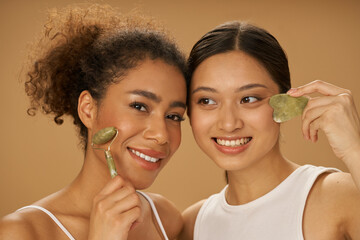 Portrait of lovely young women smiling while using jade roller and facial gua sha, posing together isolated over beige background