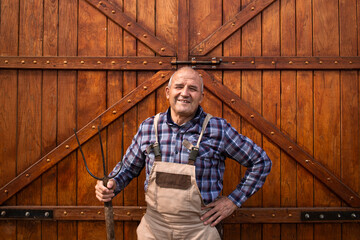 Wall Mural - Portrait of smiling hardworking farmer holding pitchfork tool and standing by wooden barn or food granary doors at domestic animals farm.