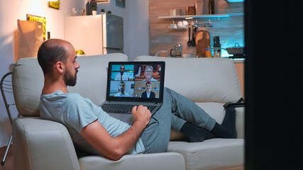 Poster - Business man in pajams lying on sofa while chatting with friends during videocall meeting using laptop computer. Caucasian male using modern technology wireless late at night in kitchen
