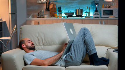 Poster - Man falling asleep in fron of TV while working on the laptop. Cozy man lying down on sofa while typing on laptop computer for social media project. Exhausted man working