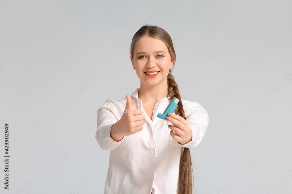 Young woman with inhaler showing thumb-up on grey background