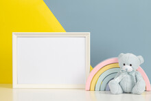 White Blank Frame With Teddy Bear And Pastel Toy Rainbow On White Desk. Baby Room Art Frame Mock Up With Baby Kid Toys Over Yellow And Gray Background