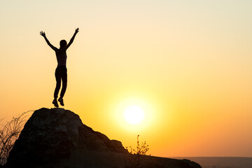 Wall Mural - Silhouette of woman hiker jumping alone on empty rock at sunset in mountains. Female tourist raising her hands up standing on cliff in evening nature. Tourism, traveling and healthy lifestyle concept.